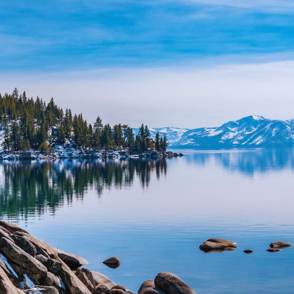 rocky shore of lake tahoe with snowy mountains and trees in the distance over the water